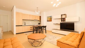 A modern living room with a sofa, a dining table with chairs, a kitchen area with cabinets and a TV. - SanPietro Vacation Apartment 050 (Plazhi San Pietro)