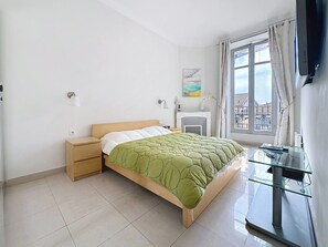 Ein Schlafzimmer mit einem Bett, Nachttischen, einem Fernseher, einem Fenster mit Vorhängen und Blick auf Gebäude.