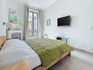 Ein Schlafzimmer mit einem Bett, einem Fernseher, einem Fenster mit Vorhängen, einem Bild und einer Heizung.