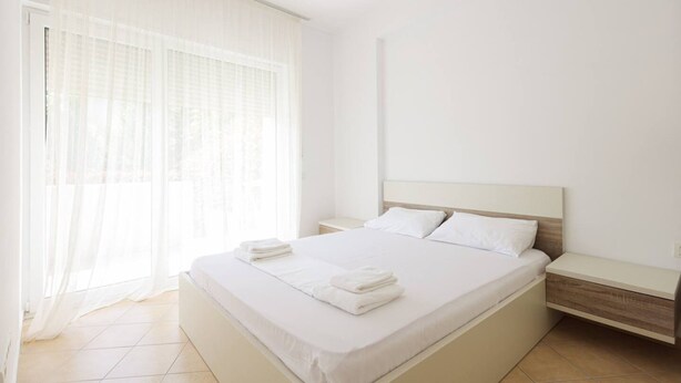 A bedroom with a bed, a nightstand, a window with sheer curtains, and a ceiling light. - Lura Apartment 05 in Plazhi San Pietro (Plazhi San Pietro)