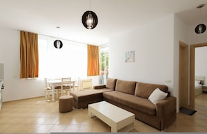 Ein modernes Wohnzimmer mit Sofa, Hocker und Couchtisch, ein Essbereich mit Stühlen sowie ein großes Fenster mit Vorhängen.