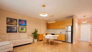 Ein modernes Wohnzimmer mit Sofa, Esstisch und Stühlen, eine Küche mit Holzschränken und einem Kühlschrank sowie eine Topfpflanze.