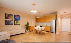 A modern living room with a sofa, dining table, and chairs, a kitchen with wooden cabinets and a refrigerator, and a potted plant. - SanPietro Vacation Apartment 053 (Plazhi San Pietro)