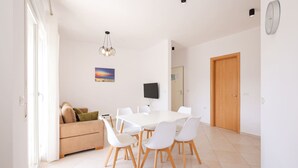 A modern dining area with a white table and chairs, a sofa with cushions, a painting on the wall, and a door. - Sunny Apartment, Balcony & Beach Access (Plazhi San Pietro)