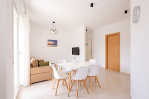 A modern dining area with a white table and chairs, a sofa with cushions, a painting on the wall, and a door. - Sunny Apartment, Balcony & Beach Access (Plazhi San Pietro)