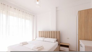 Un dormitorio moderno con cama, mesitas de noche, un armario y una ventana con cortinas traslúcidas.