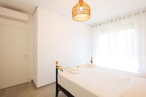 A bedroom with a bed, a window with curtains, a door, and a hanging light fixture. - Boho-Chic Apartment with Garden Views (Plazhi San Pietro)