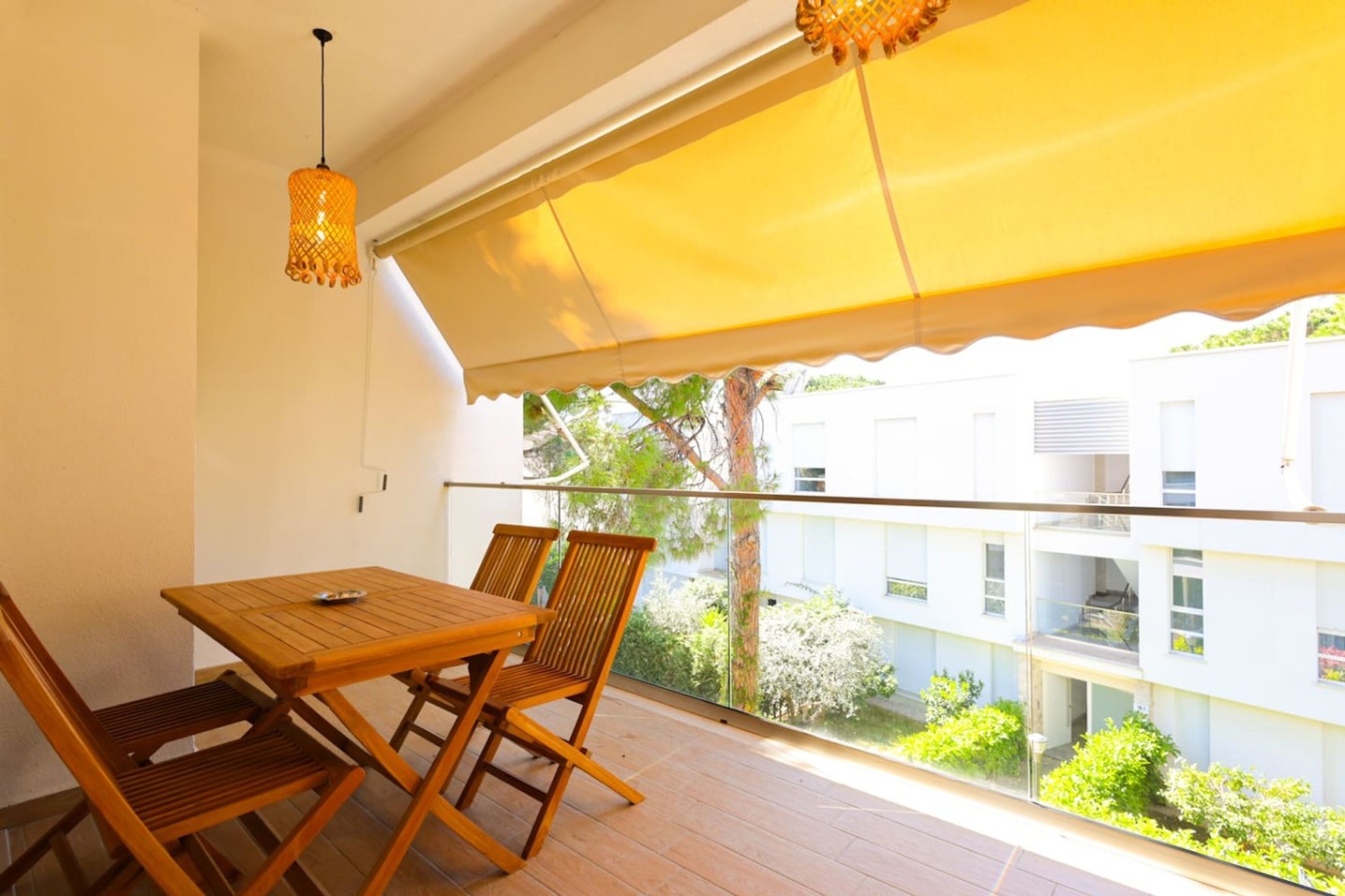 A balcony with a wooden table and chairs, a yellow awning, and a view of buildings and greenery.