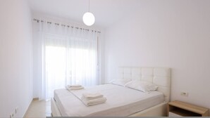 A bedroom with a bed, white curtains, a bedside table, and a hanging light fixture. - Lura Apartment 04 in Plazhi San Pietro (Plazhi San Pietro)