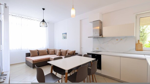 A modern kitchen with a dining area, a sofa, and a window with blinds. - Lura Apartment 04 in Plazhi San Pietro (Plazhi San Pietro)