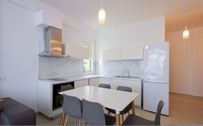 A modern kitchen with white cabinets, a stainless steel range hood, a built-in oven, a refrigerator, and a dining area with a table and chairs. - Lura Apartment 04 in Plazhi San Pietro (Plazhi San Pietro)
