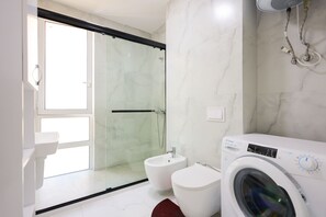 A modern bathroom with a glass shower enclosure, a toilet, a bidet, and a washing machine. - SanPietro Vacation Apartment 051 (Plazhi San Pietro)