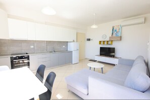 Una cocina moderna con armarios blancos, horno empotrado y refrigerador. Cuenta con un área de comedor con mesa y sillas blancas, y un sofá de gris claro. Un televisor de pantalla plana está montado en la pared.