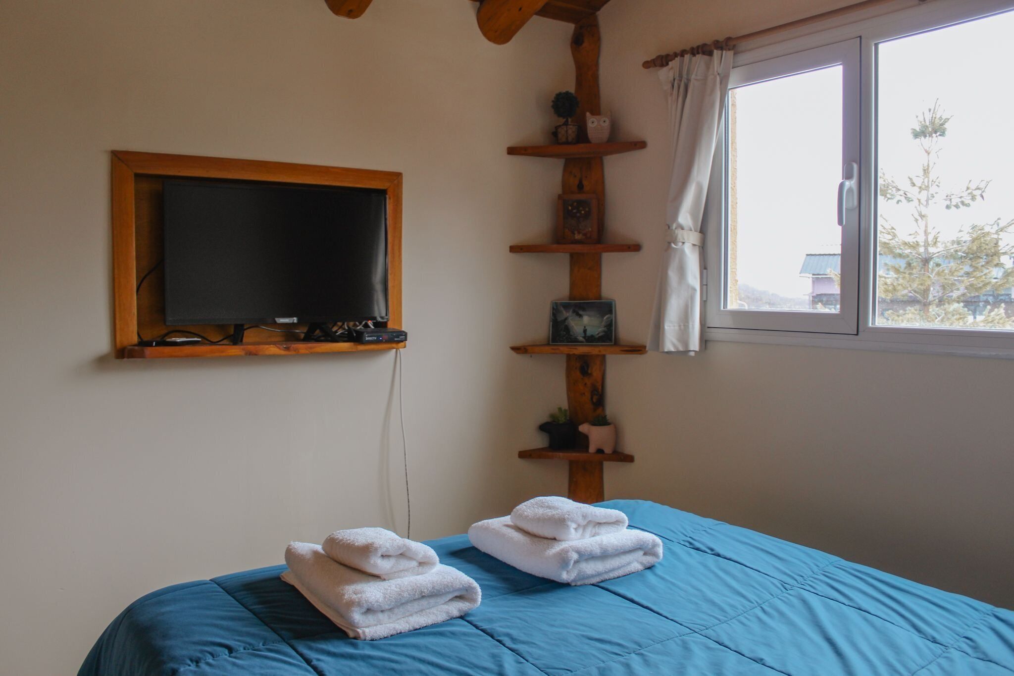 Una habitación con una cama, televisión montada en la pared, una ventana con cortinas y una estantería con diversos objetos.