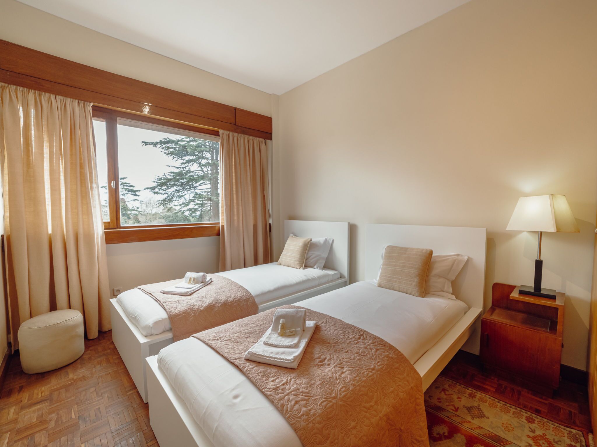 A hotel room with two beds, a window with curtains, a bedside lamp, and a small table.