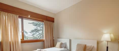 A hotel room with two beds, a window with curtains, a bedside lamp, and a small table.