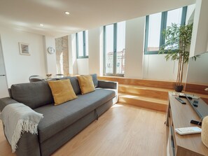 Ein modernes Wohnzimmer mit grauem Sofa, Holzboden, großen Fenstern und einer Pflanze.