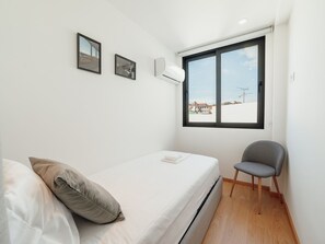 Una habitación pequeña con cama individual, ventana, una silla y dos unidades de aire acondicionado fijadas a la pared.