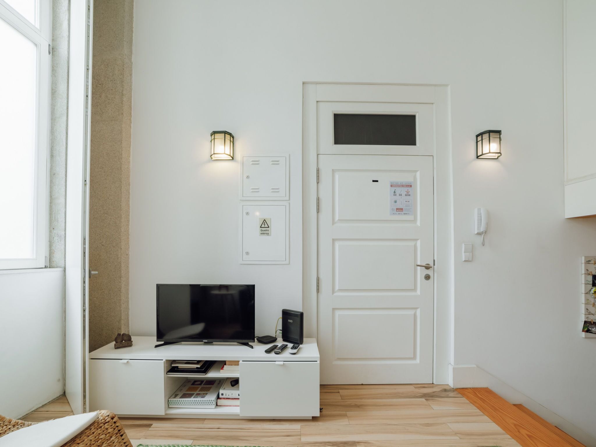 A room with a white door, a flat-screen TV on a white cabinet, two wall-mounted light fixtures, and a small shelf with books and a remote control.