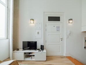 Una habitación con una puerta blanca, un televisor de pantalla plana sobre un mueble blanco, dos lámparas empotradas en la pared y un estante pequeño con libros y un control remoto.