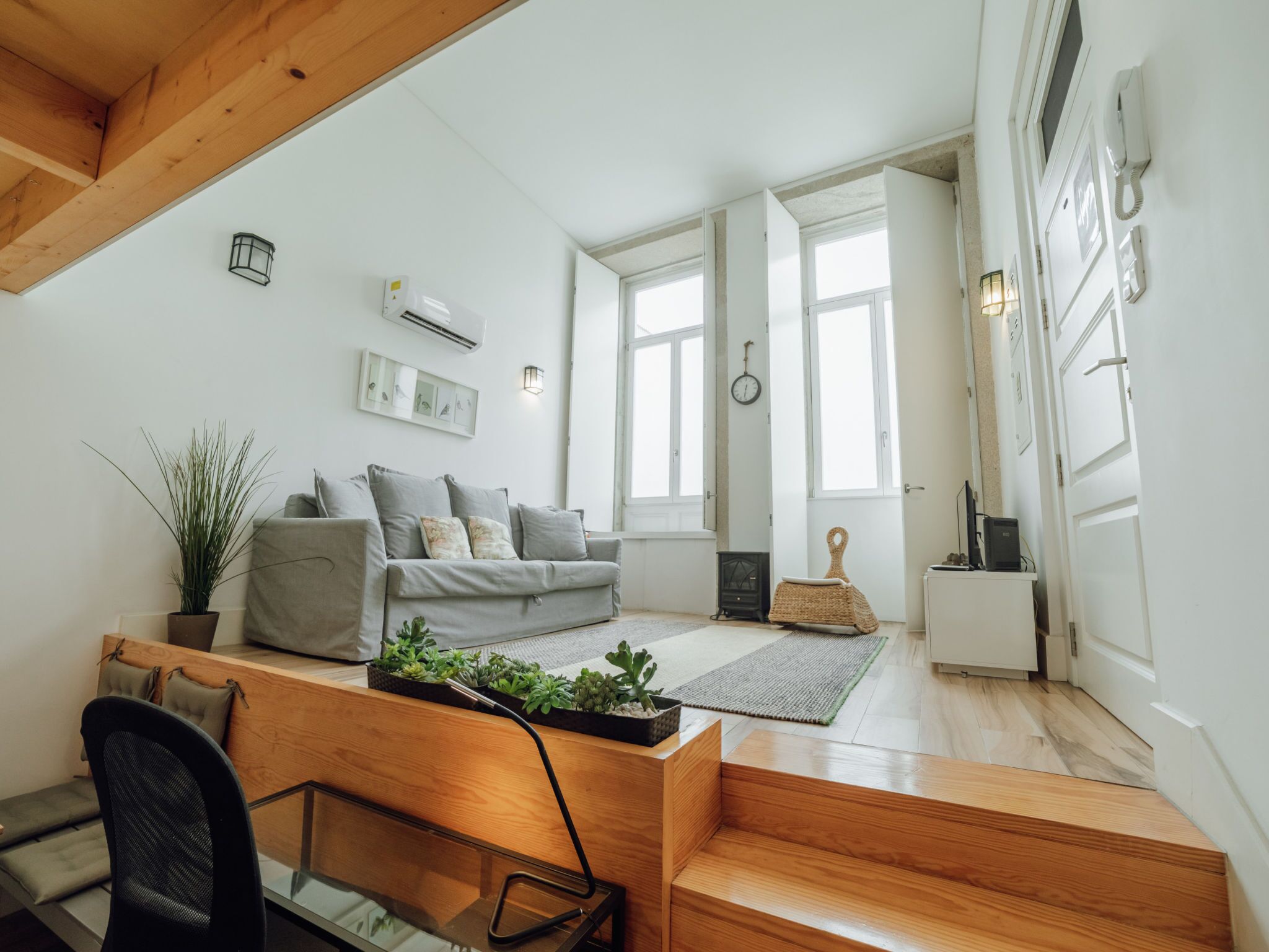 A modern living room with a grey sofa, a wooden coffee table with plants, a staircase, and a large window providing natural light.
