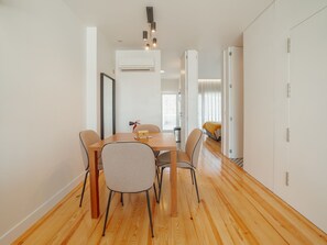 Uma área de refeições moderna com uma mesa e cadeiras de madeira, um aparelho de ar condicionado acima e vista para um quarto com uma cama e cortinas.