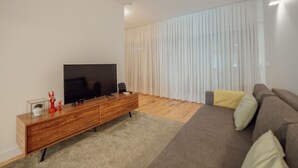 A modern living room with a flat-screen TV on a wooden console, a sofa with cushions, and a large window with sheer curtains. - Apartment  by Interhome (Porto)