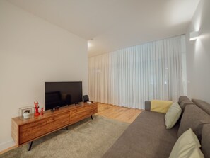 Uma sala de estar moderna com uma televisão de tela plana sobre um console de madeira, um sofá com almofadas e uma grande janela com cortinas leves.