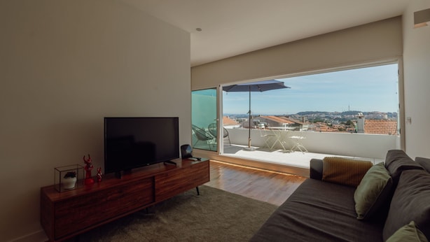 A modern living room with a flat-screen TV on a wooden console, a large window with a view of a cityscape, and a balcony with outdoor furniture. - Apartment  by Interhome (Porto)