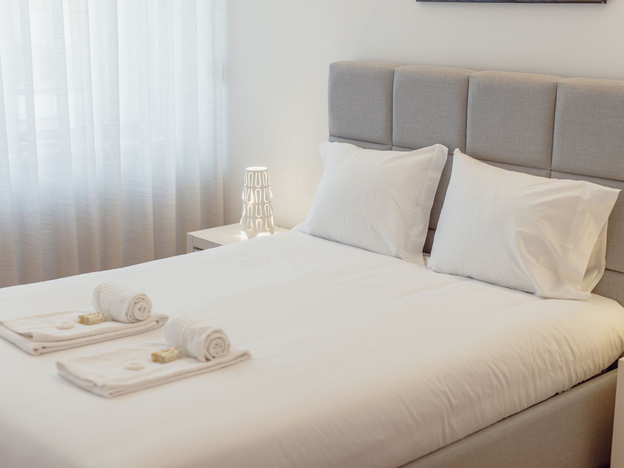 Un lit bien fait, avec du linge de lit blanc et des oreillers, une table de chevet avec une lampe et une fenêtre ornée de rideaux légers.