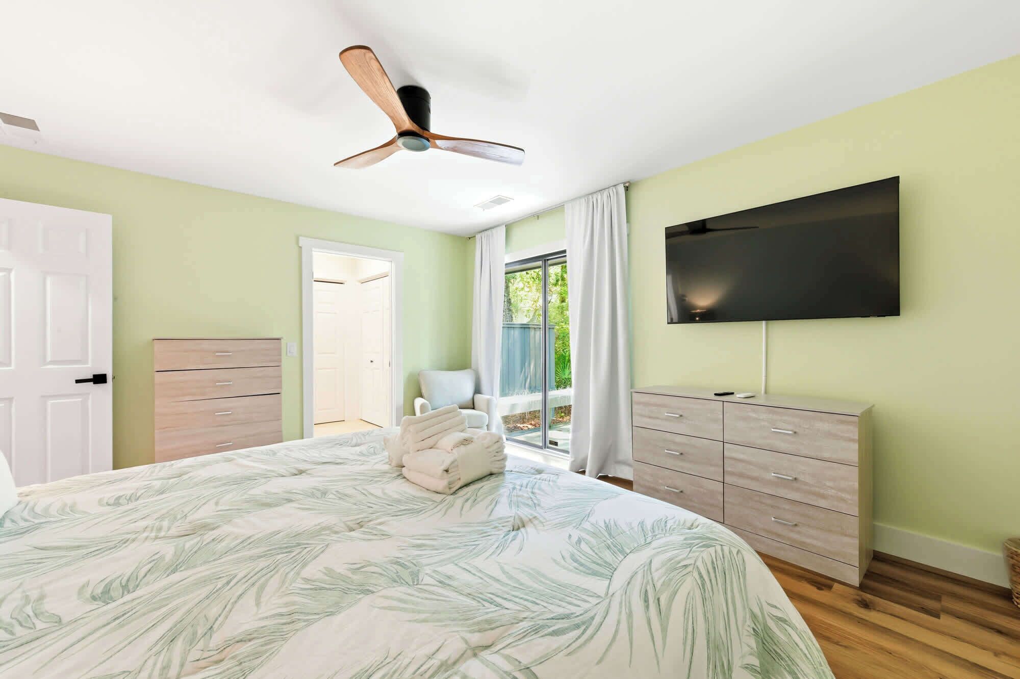 Un dormitorio con una cama, un ventilador de techo, un televisor, un aparador y una puerta que da a un balcón.