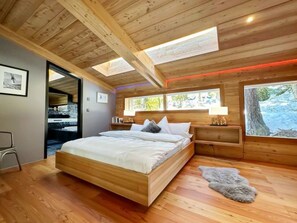 Ein Schlafzimmer mit Holzboden, einem Bett, einem Nachttisch, einer Lampe, einem Fenster mit Blick und einer Tür zu einem anderen Raum.