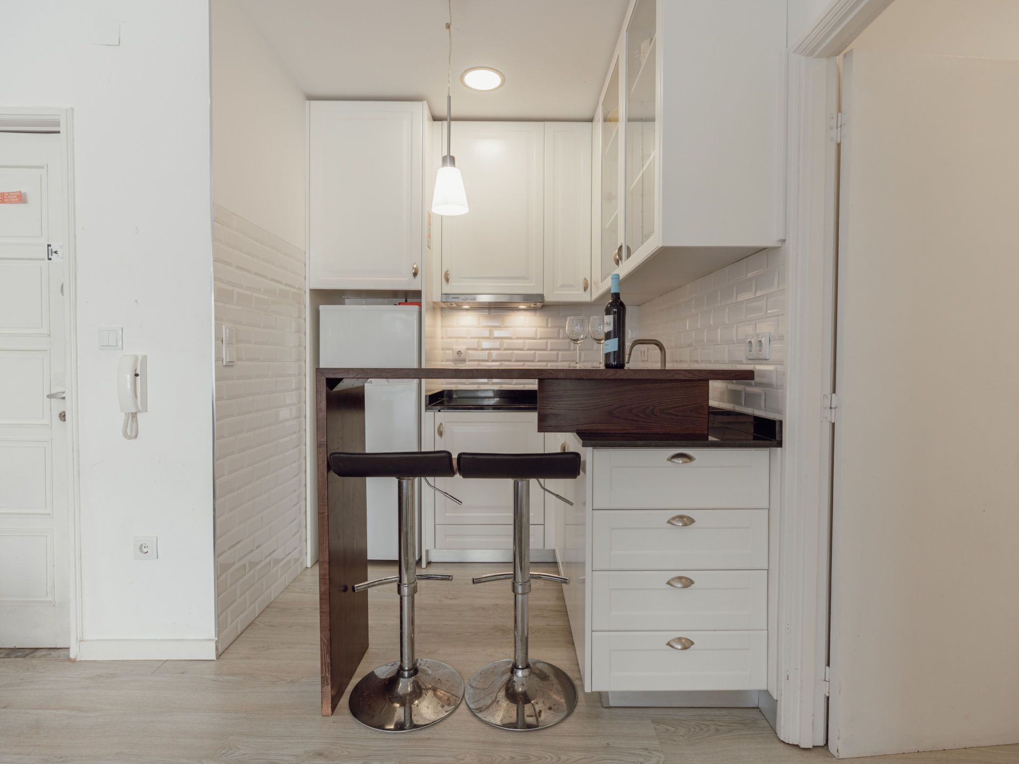 A compact kitchen with white cabinetry, a wooden countertop, and two bar stools.