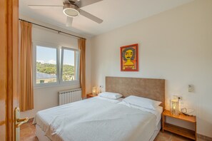 Un dormitorio con cama, mesita de noche, ventilador de techo, ventana con cortinas e iluminación de pared.