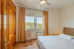 A bedroom with a bed, a chair, a window with curtains, a radiator, and a ceiling fan.