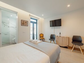 Ein Hotelzimmer mit einem Bett, einem Flachbildfernseher, zwei Stühlen, einem kleinen Holfnachtkästchen und einer Duschkabine aus Glas.