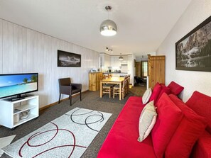 Ein Wohnzimmer mit rotem Sofa, einem Fernseher auf einem Schrank, einem Essbereich mit Tisch und Stühlen sowie einer Küche im Hintergrund.