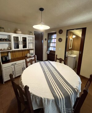 Un coin repas avec une table ronde, des chaises, une cuisine équipée d’armoires et d’un appareil à café, ainsi qu’un luminaire suspendu.