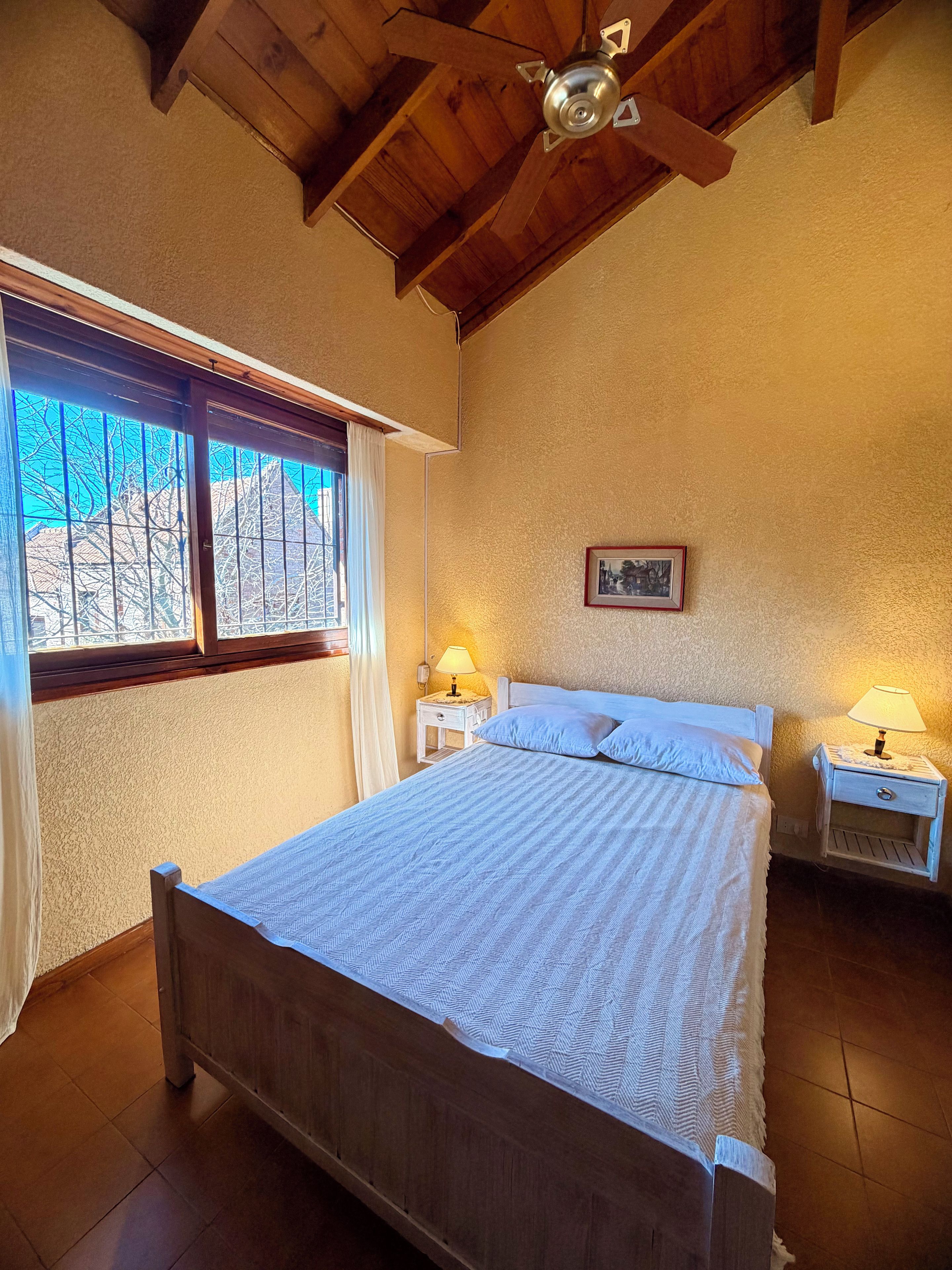 A bedroom with a wooden bed, two bedside tables with lamps, a window with curtains, and a ceiling fan.