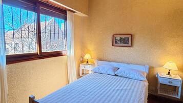 A bedroom with a wooden bed, two bedside tables with lamps, a window with curtains, and a ceiling fan.