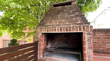 A brick-built outdoor oven with a metal grill, surrounded by a wooden fence and a tree.