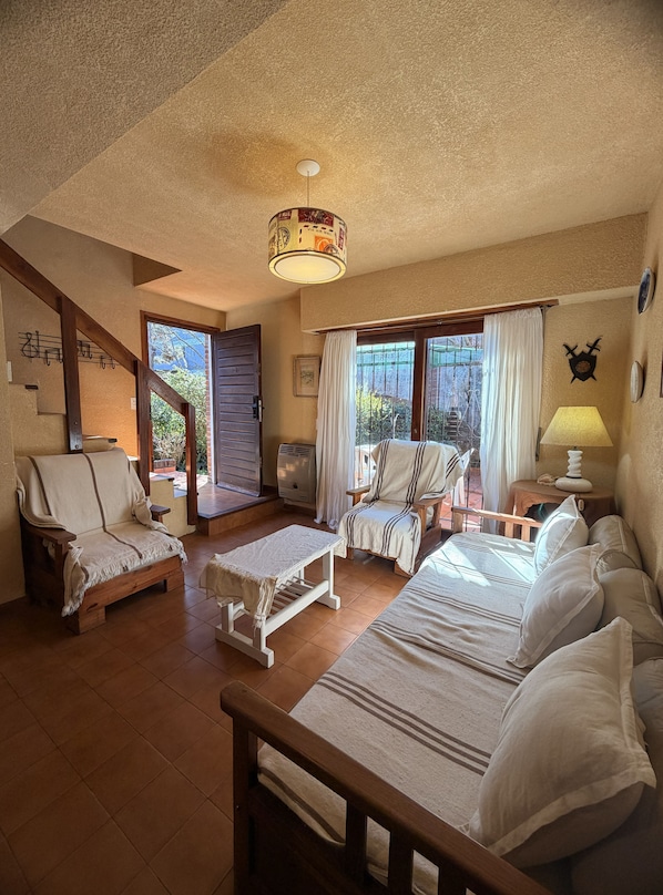 A cozy room with a bed, a sofa, a chair, and a table. There is a window with curtains, a ceiling light, and a wall-mounted decoration.