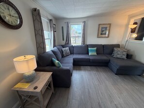 Ein modernes Wohnzimmer mit einem Ecksofa, einem kleinen Holztisch mit einer Lampe und einer großen Wanduhr.