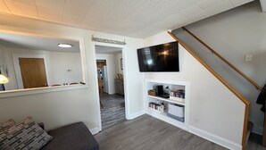 Uma sala de estar com um sofá cinzento, uma TV fixada na parede e uma escada.