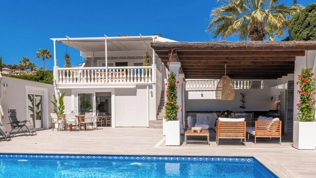 A poolside area with a clear blue pool, a covered seating area with wooden loungers, and a white building with a balcony. - Villa Lorenza by Interhome (Moraira)