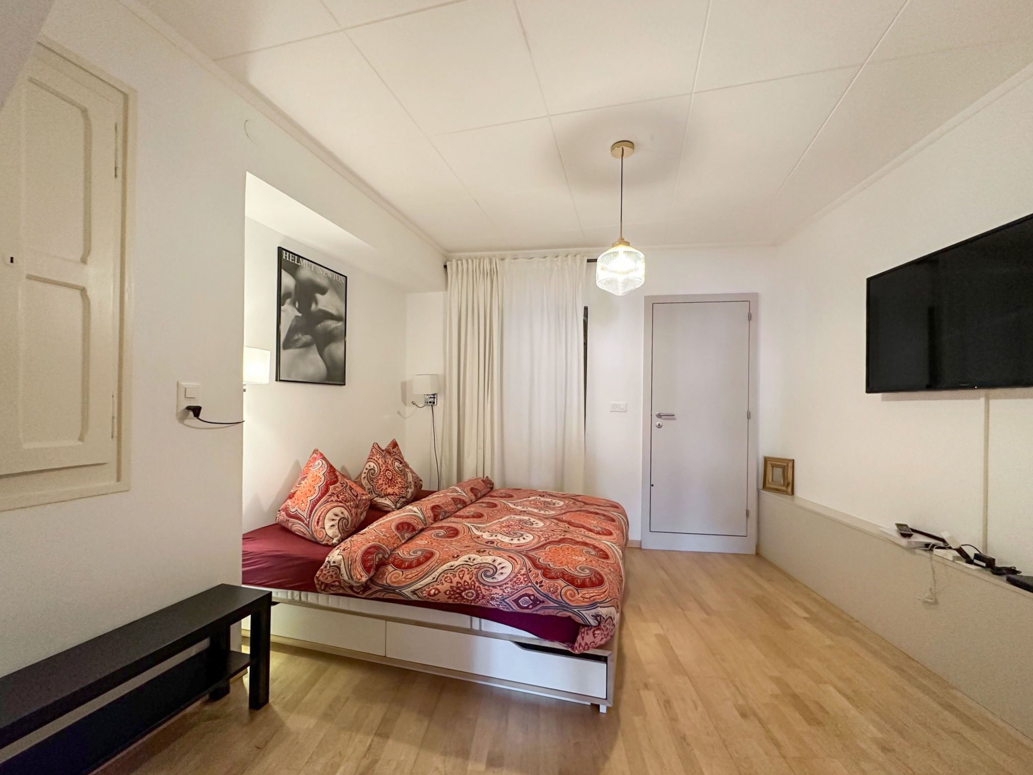 Een slaapkamer met een bed, een bankje, een deur, een aan de muur bevestigde televisie en een plafondlamp.