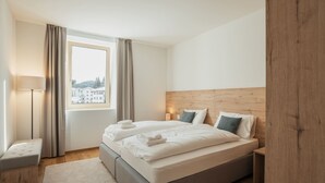 Una habitación de hotel moderna con una cama grande, una mesita de noche, una lámpara y vistas a edificios a través del ventanal.