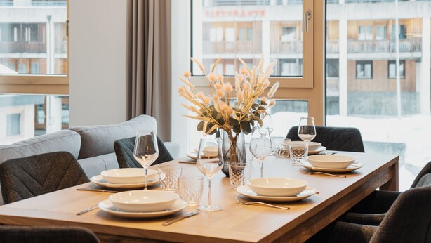 Un coin repas avec une table en bois dressée pour quatre personnes, un vase contenant des fleurs séchées et une vue sur un immeuble doté de balcons.