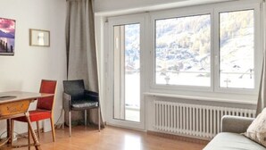 A room with a wooden floor, a dining table with chairs, a sofa, a radiator, and large windows with a view of a snowy landscape.