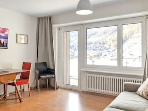 Ein Zimmer mit Holzboden, einem Esstisch mit Stühlen, einer Couch, einem Heizkörper und großen Fenstern mit Blick auf eine verschneite Landschaft.